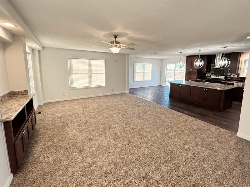 An empty living room with a ceiling fan and a kitchen in the background.