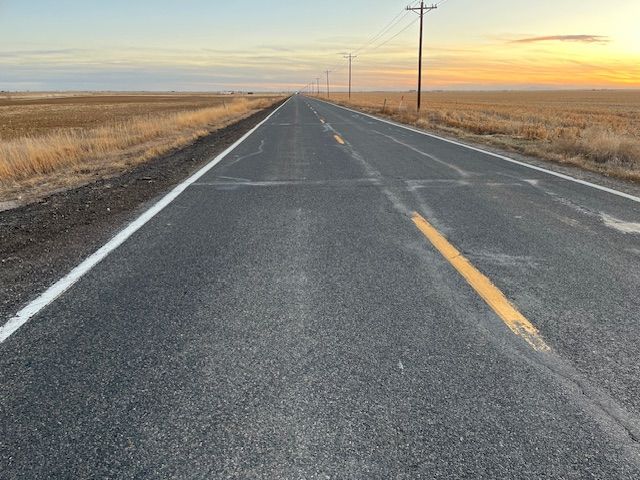 Paved road stretching into the distance under a pale sky. Fields on either side, power poles visible.