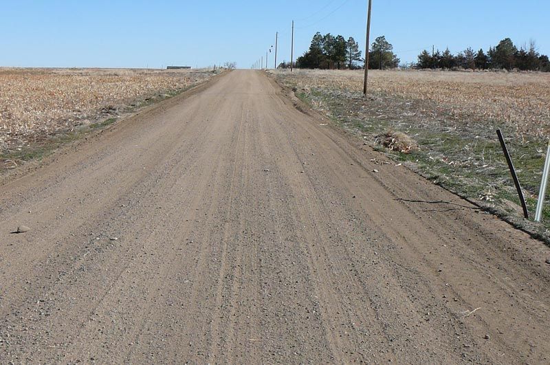 Gravel road stretches through rural landscape. Brown fields on either side, blue sky above.