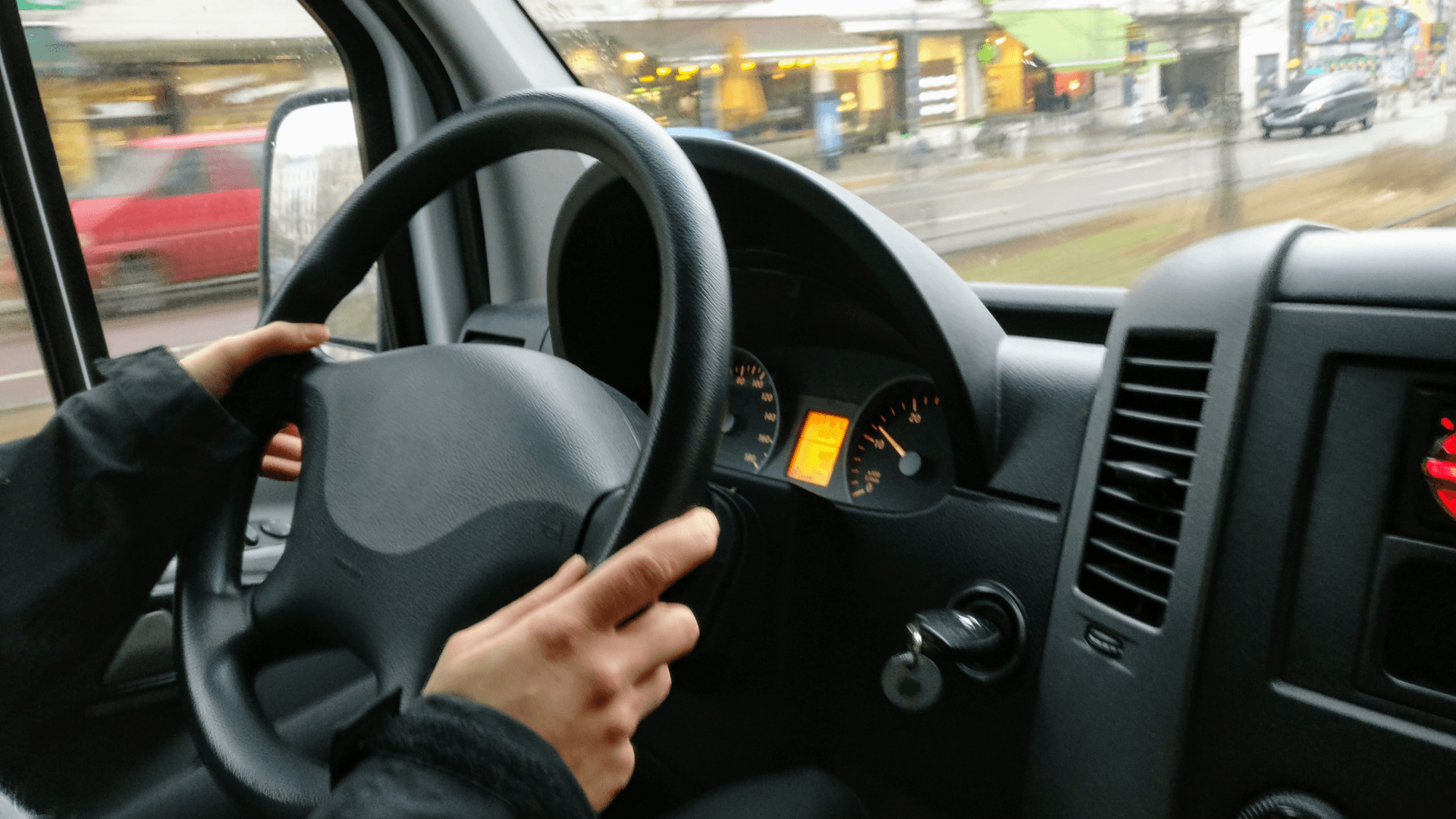 Interior driver's side of a car. The driver's hands are on the wheel as they drive through a city.