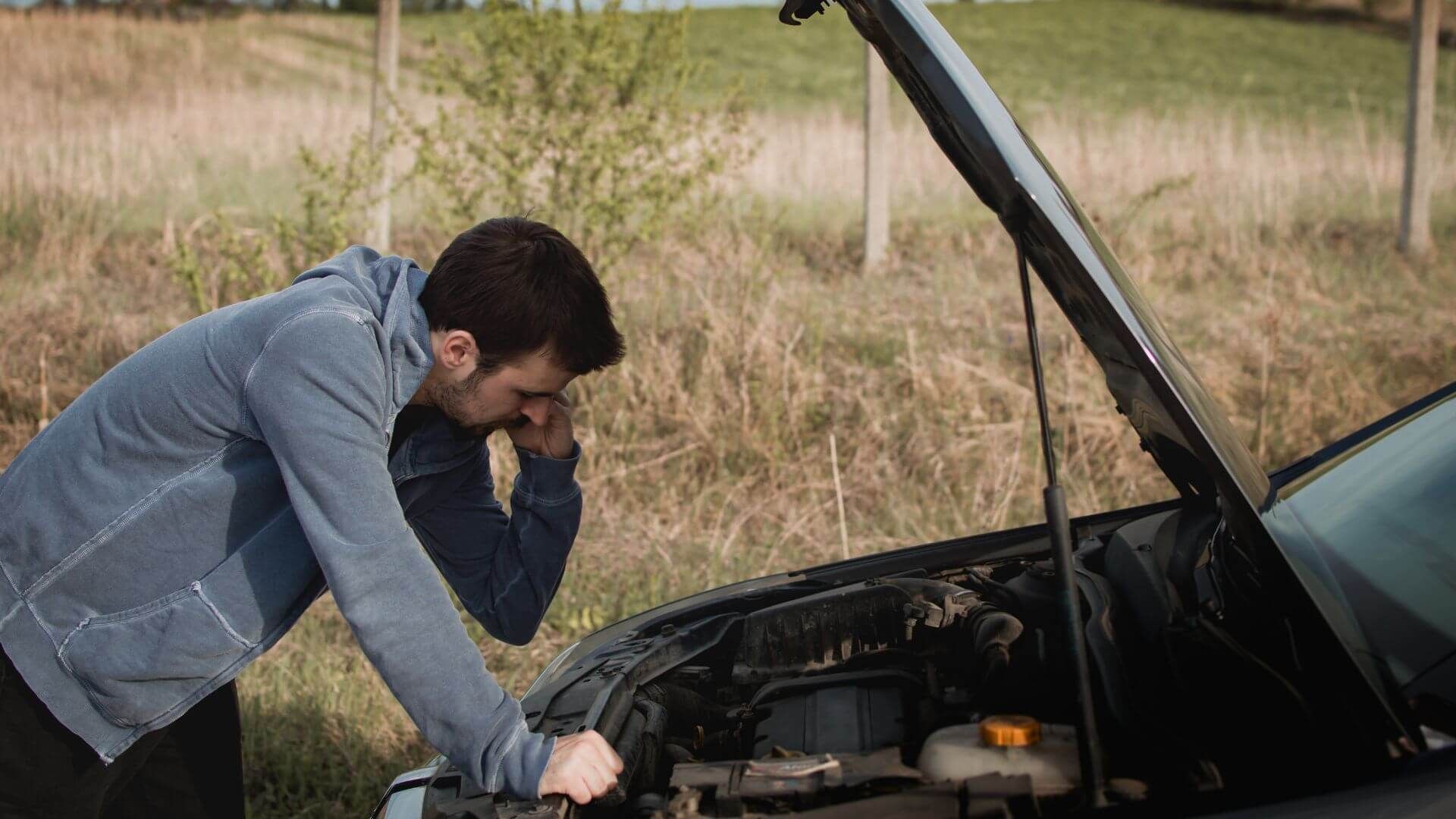 Man talking on the phone, leaning over a car engine with the hood popped on the side of the road.