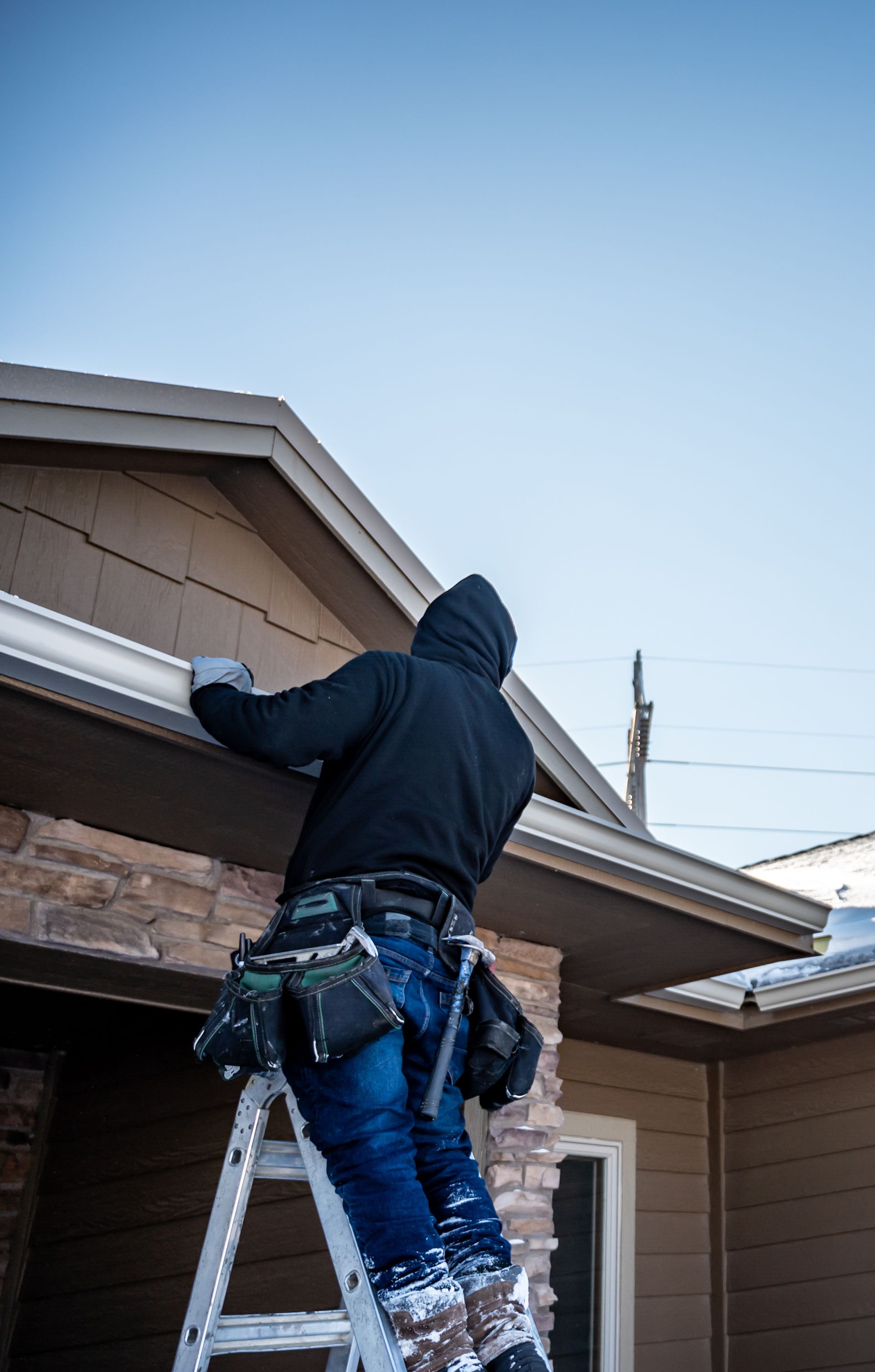 Person in a hooded sweatshirt on a ladder installing gutters on a house. Blue sky overhead.