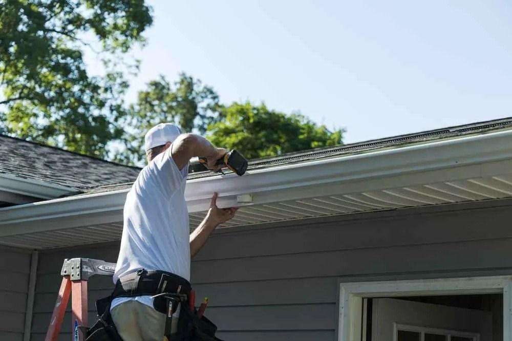 Person on ladder installing gutter on a house, blue sky and trees in background.