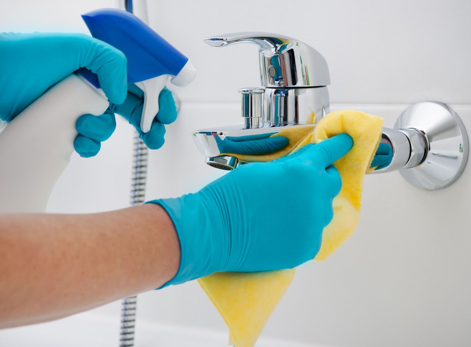 Person wearing blue gloves cleaning a chrome faucet with a yellow sponge and spray bottle.