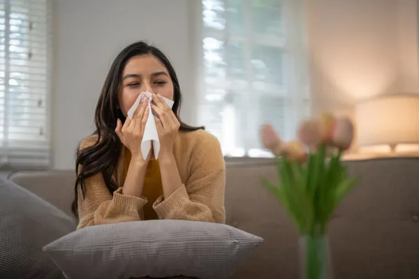 Woman indoors, blowing nose with tissue. Brown sweater, sofa, tulips, window.