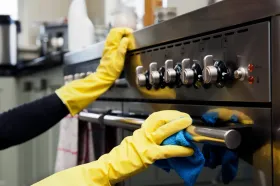 Hands in yellow gloves cleaning a stainless steel oven with a blue cloth in a kitchen.