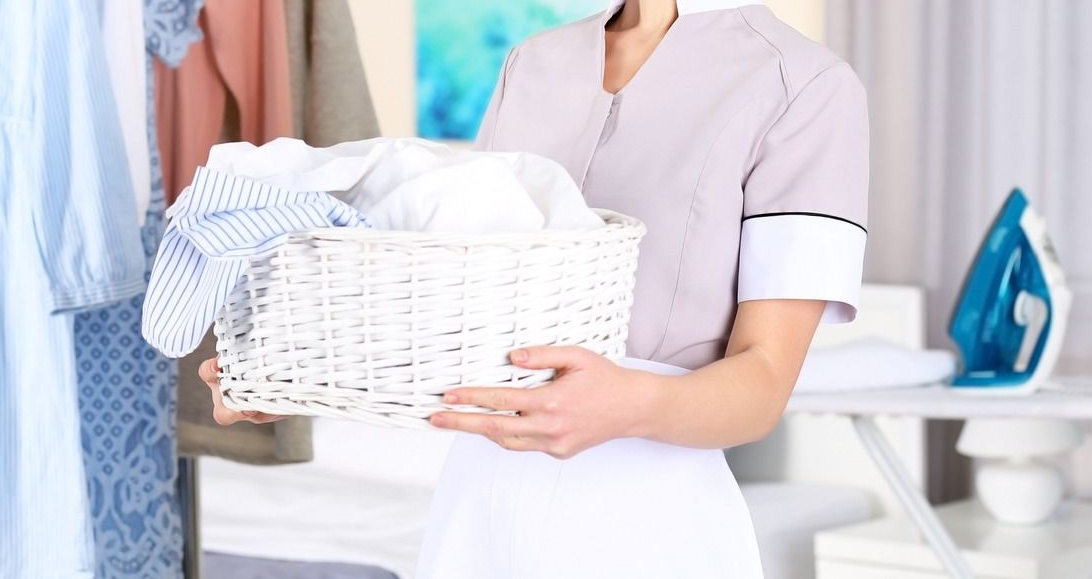Maid holding a white basket of clean laundry near an ironing board with a blue iron.
