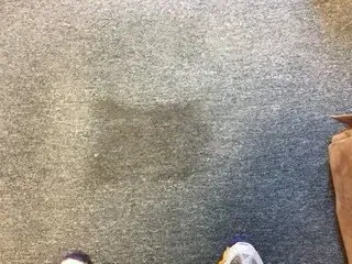 Carpeted floor with a dark square stain in the center and a brown bag at the right edge.
