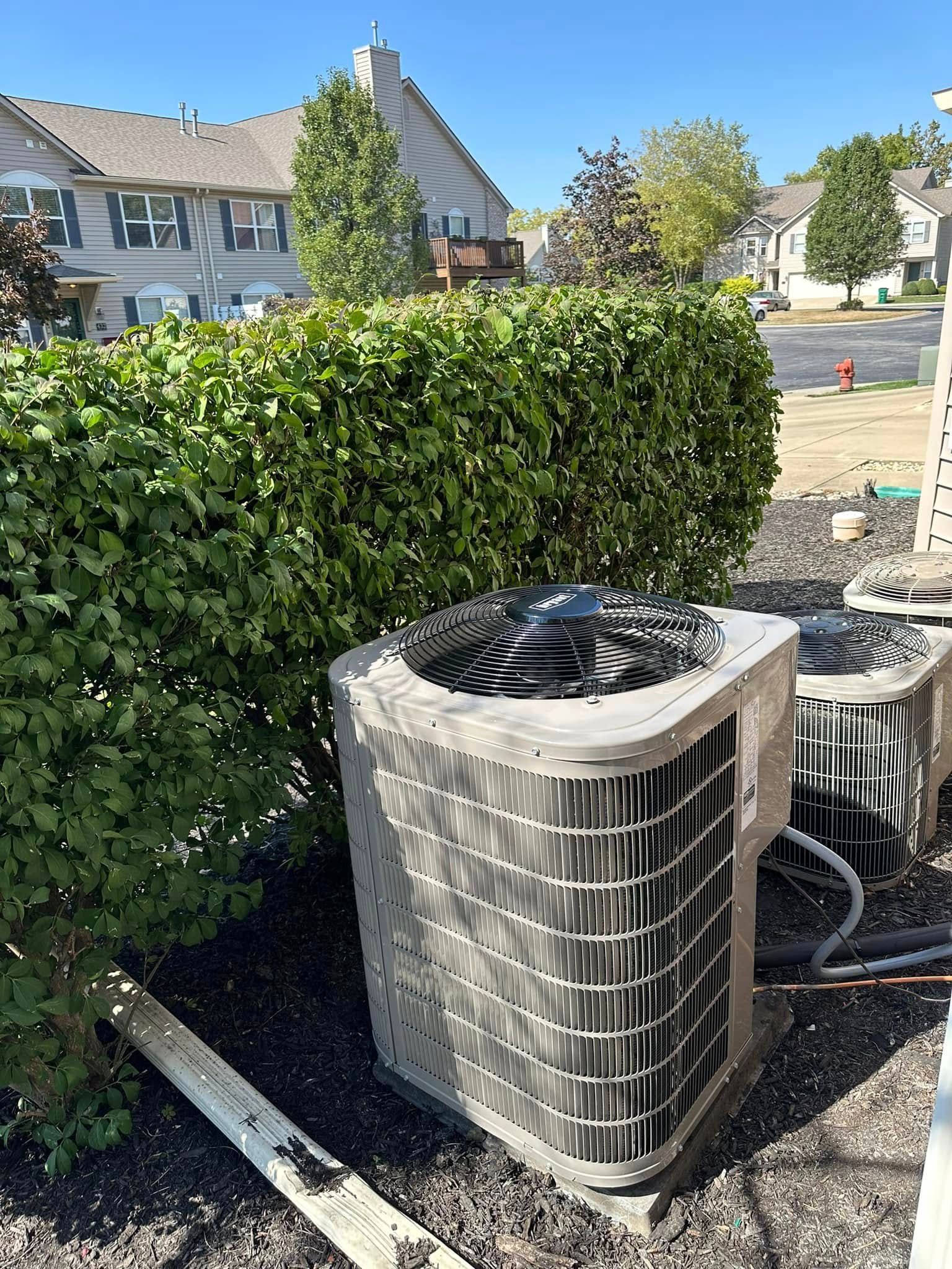 Air conditioning units next to a hedge, in front of houses on a sunny day.
