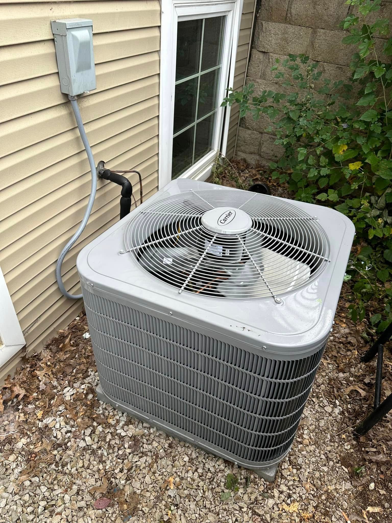 Air conditioning unit outside a building, gray with fan visible. Next to a window and bushes.