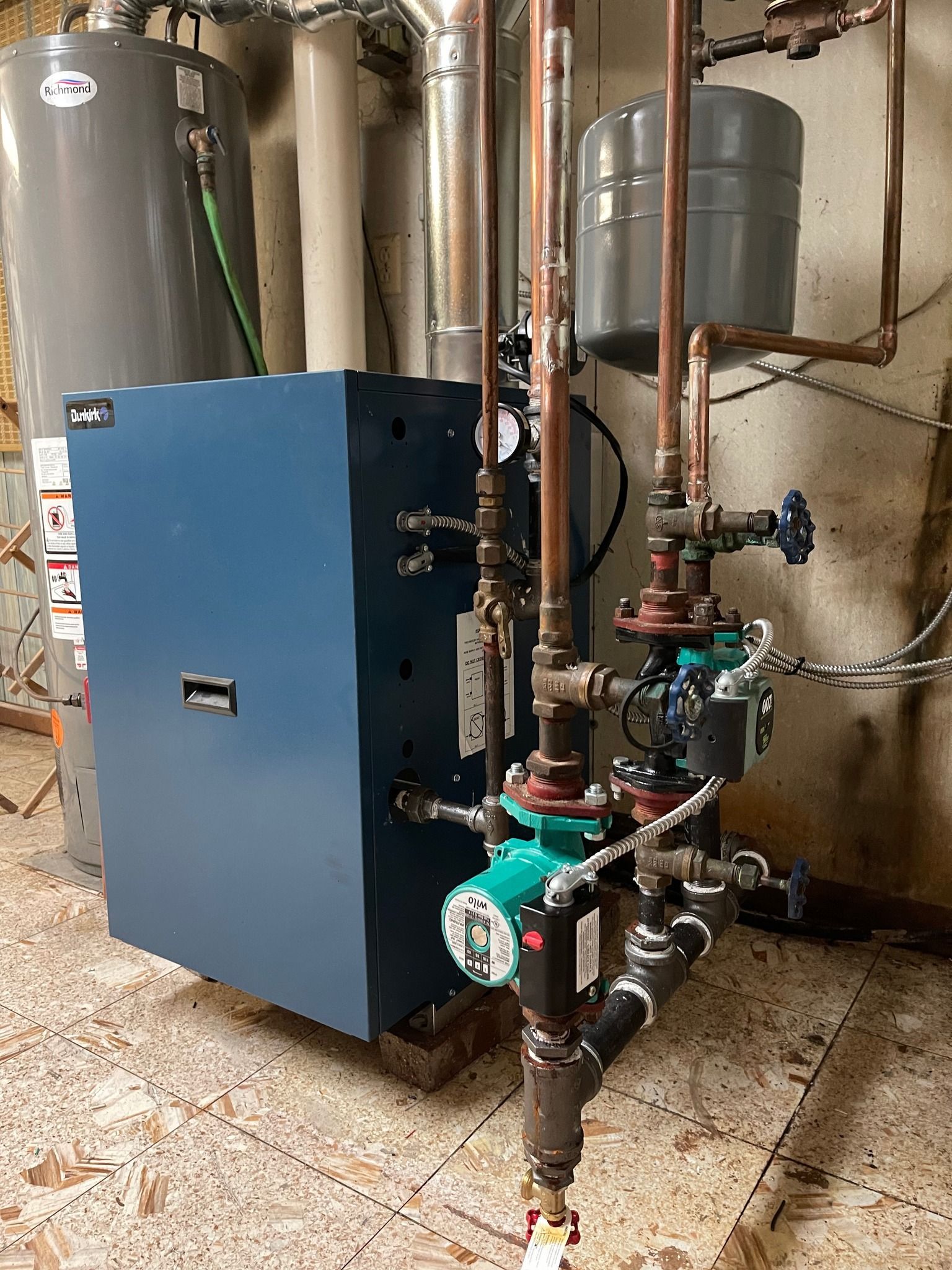 Blue boiler and associated plumbing in a utility room. Copper pipes, green pumps, grey tank.