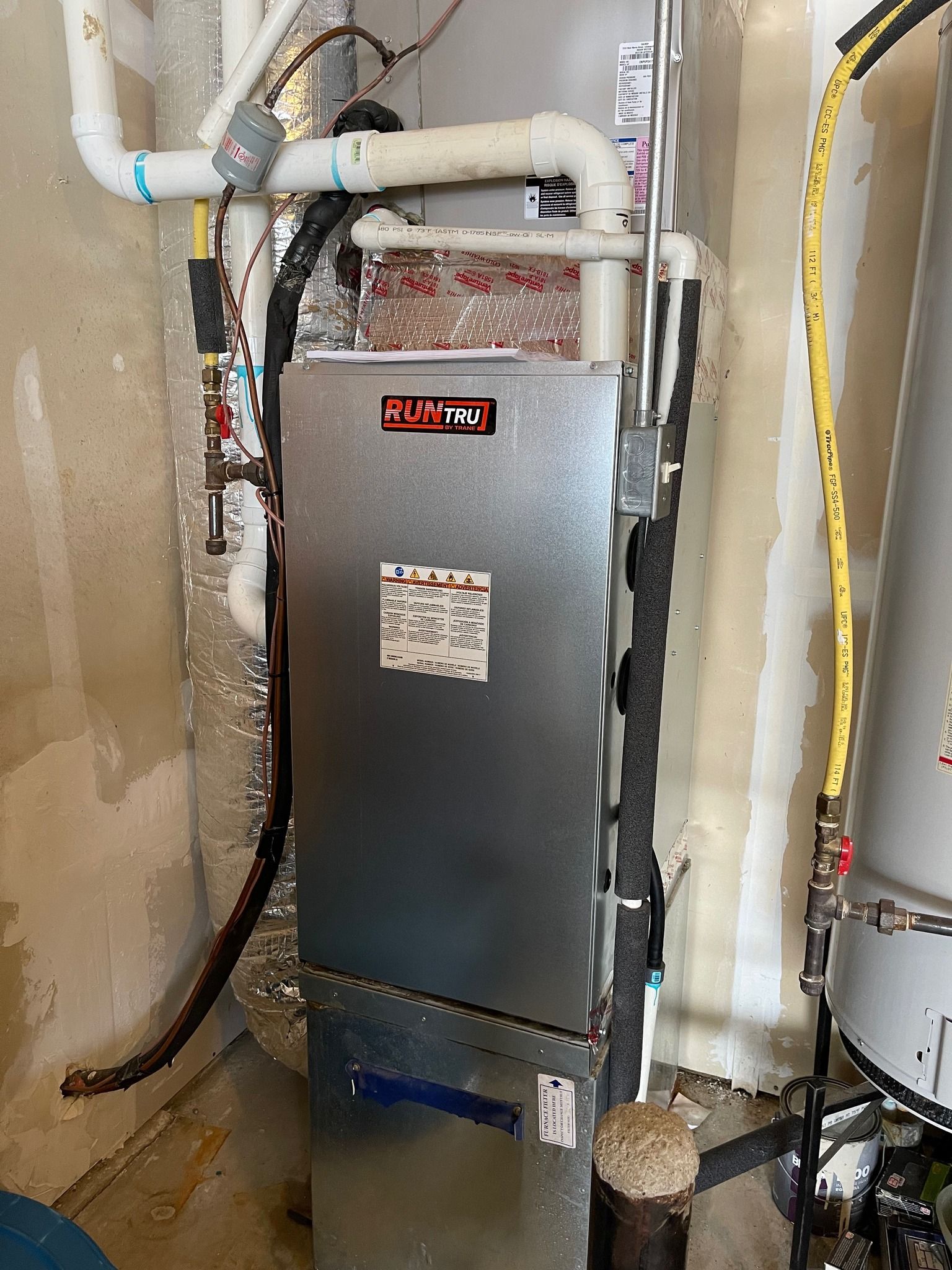 Furnace and water heater in a utility room. Gray metal furnace with red logo, pipes, and water heater on the right.