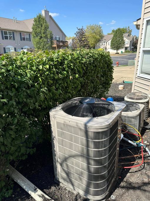 Two outdoor AC units next to a trimmed green hedge, with houses and blue sky in the background.