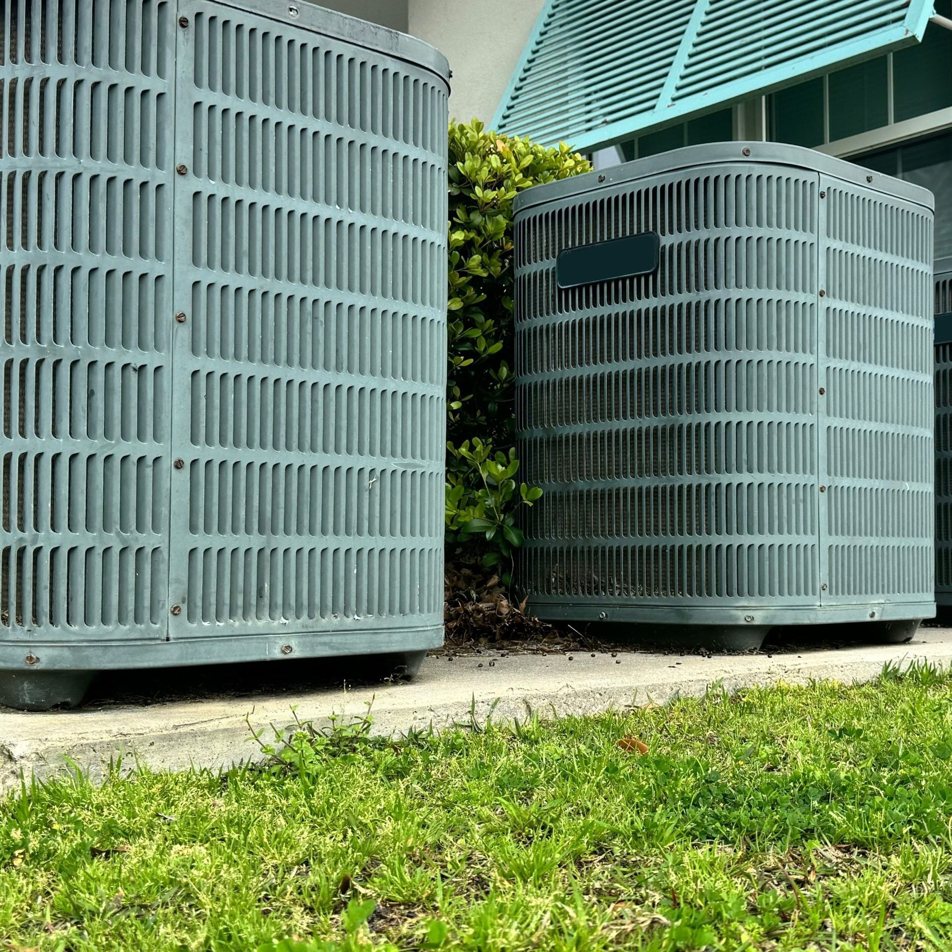 Three gray air conditioning units on concrete near grass and building.
