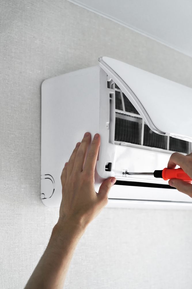 Person using a screwdriver to open a white wall-mounted air conditioner unit for maintenance or repair.