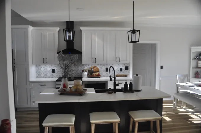 A modern kitchen with white cabinets, a dark island with three stools, a black stove hood, and white counters.