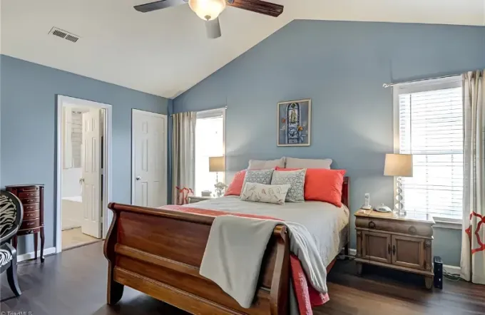 A bright, vaulted-ceiling bedroom with blue walls, a wooden sleigh bed, and two windows with white curtains.
