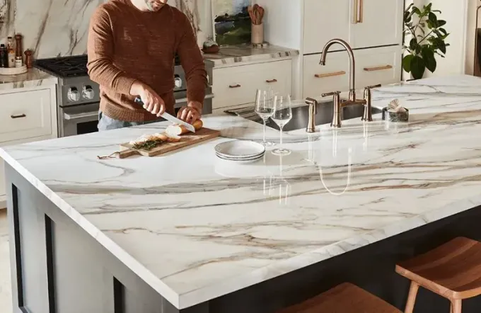 A person in a brown sweater slices bread on a marble kitchen island countertop with a sink and two wine glasses nearby.