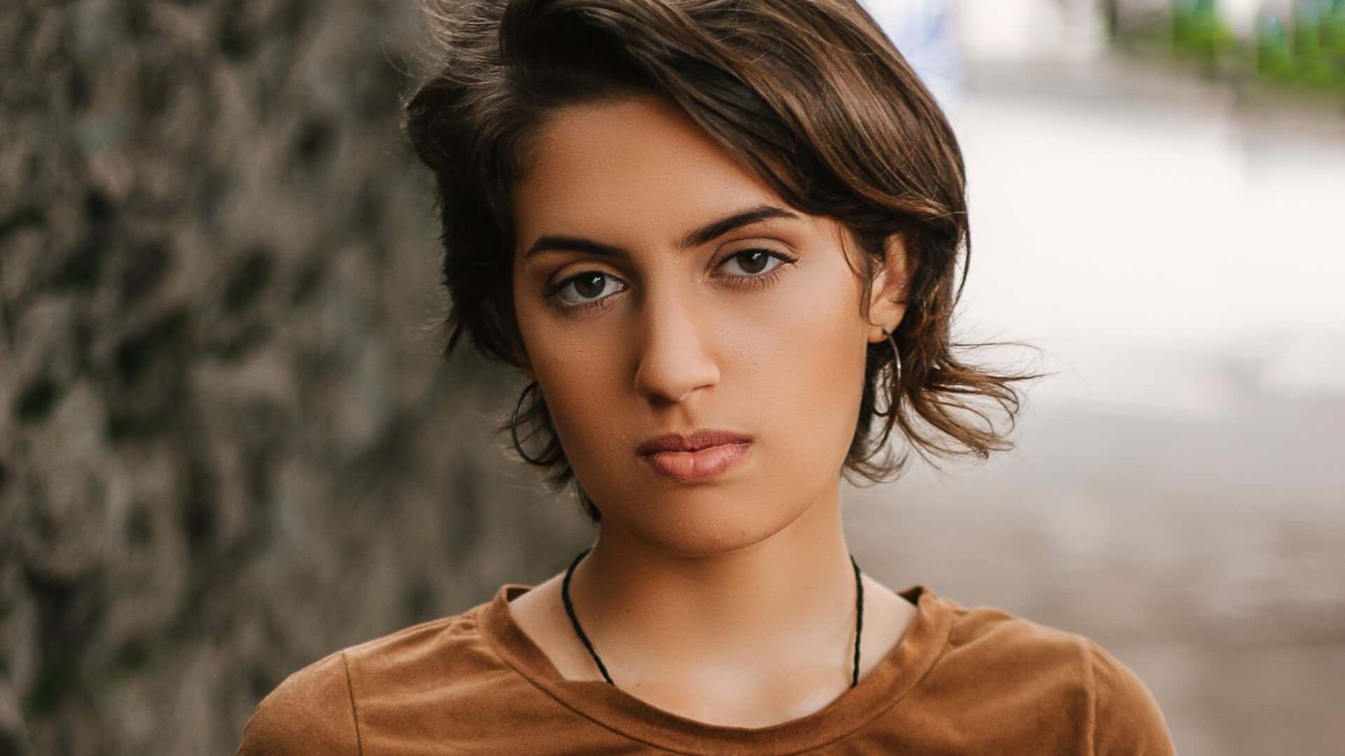 A woman in a brown t-shirt is standing on a sidewalk holding a camera.