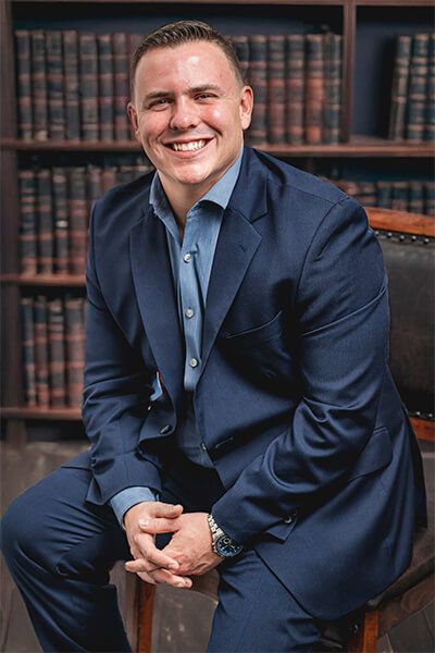 A man in a suit is sitting on a chair in front of a bookshelf.