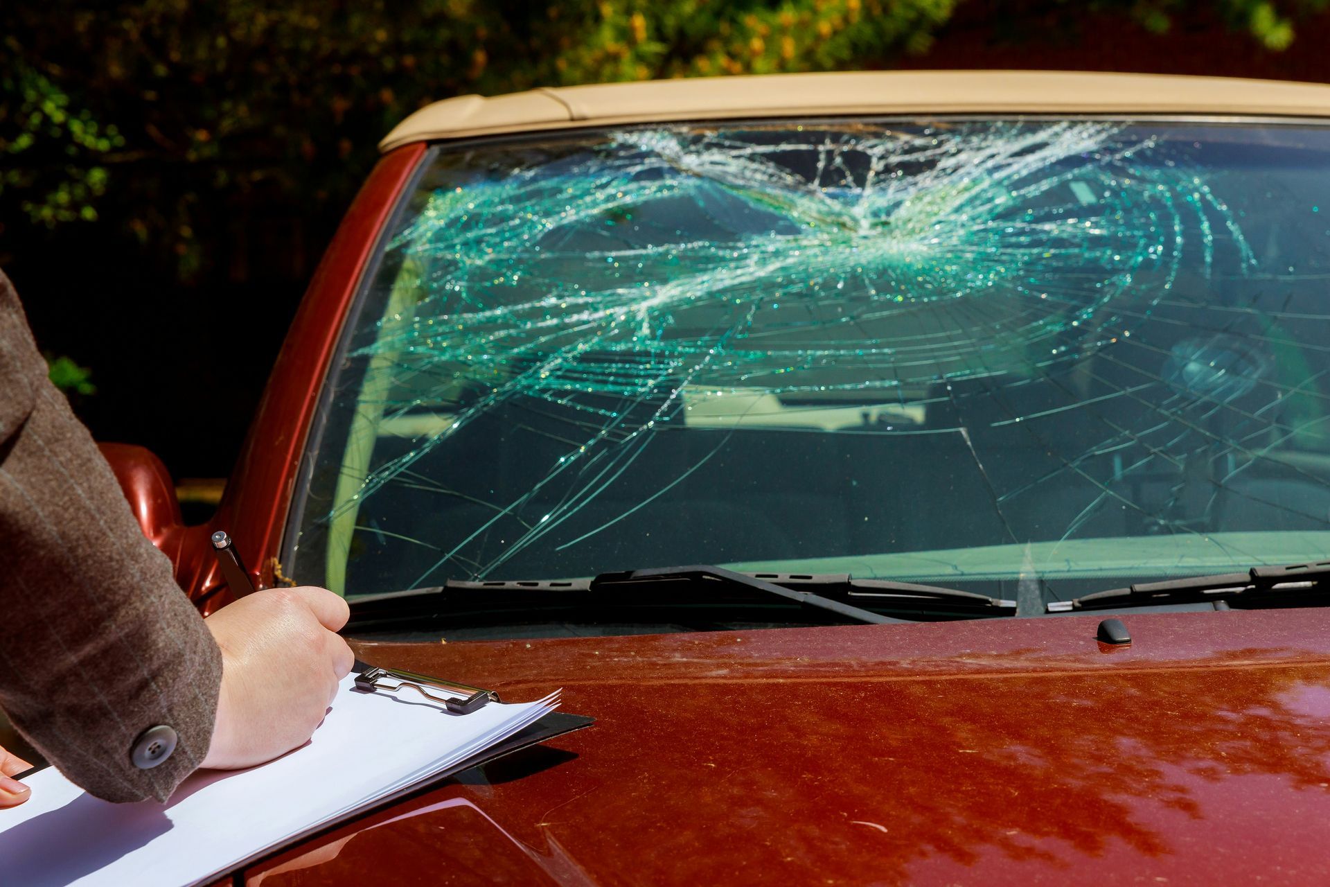 A person is writing on a clipboard next to a car with a broken windshield.