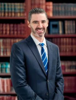 A man in a suit and tie is standing in front of a bookshelf.