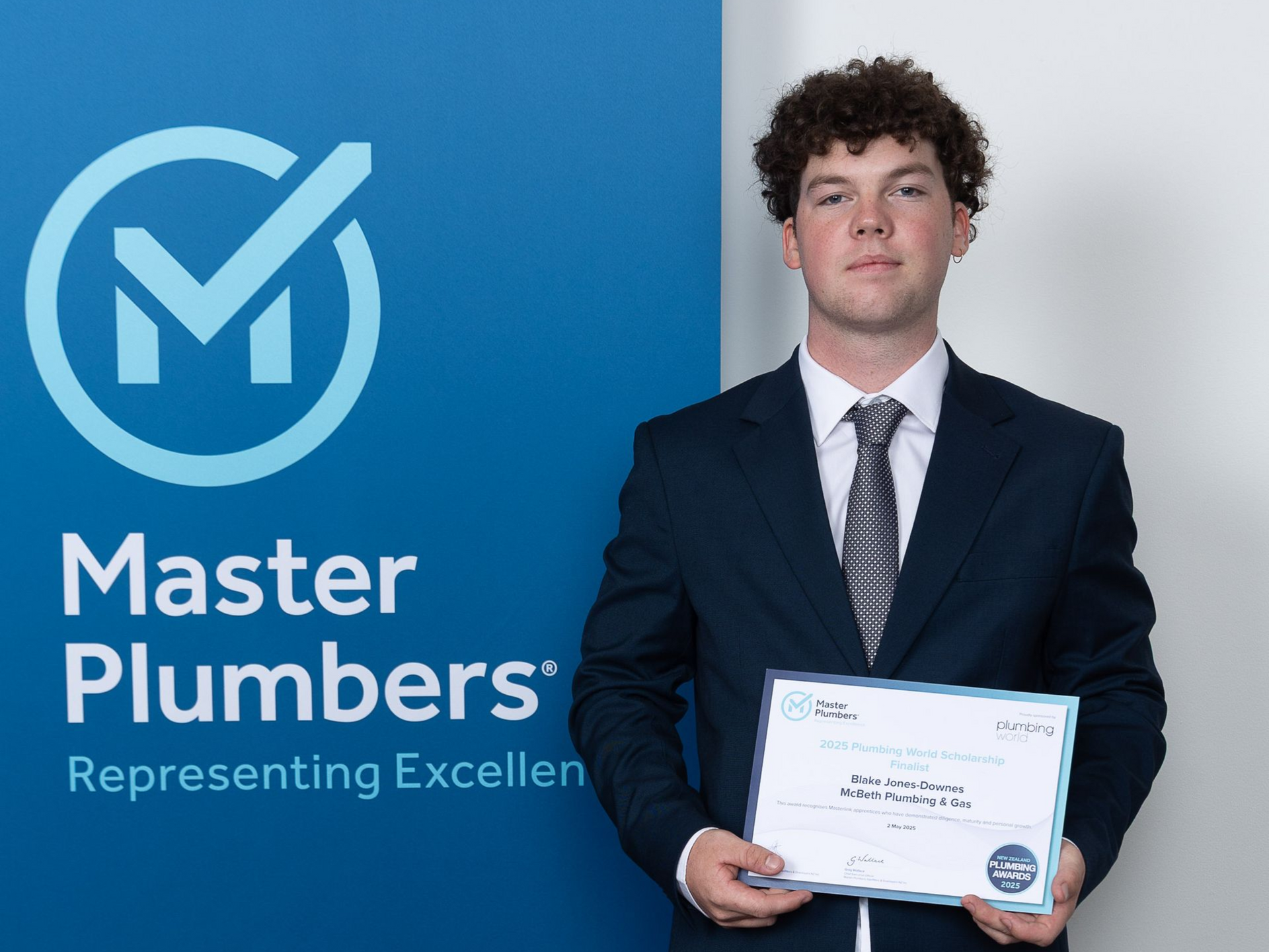 Young man in a suit holding a certificate, standing in front of a blue Master Plumbers banner.