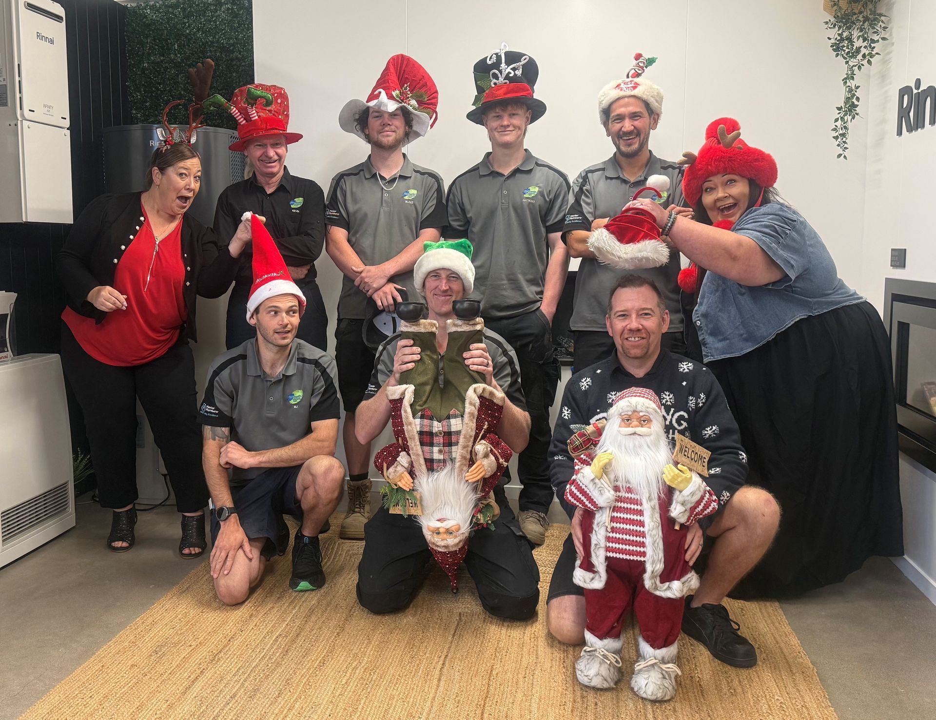 Group of people posing, some wearing Christmas attire. Outdoors, in front of a building, bright. Smiles.