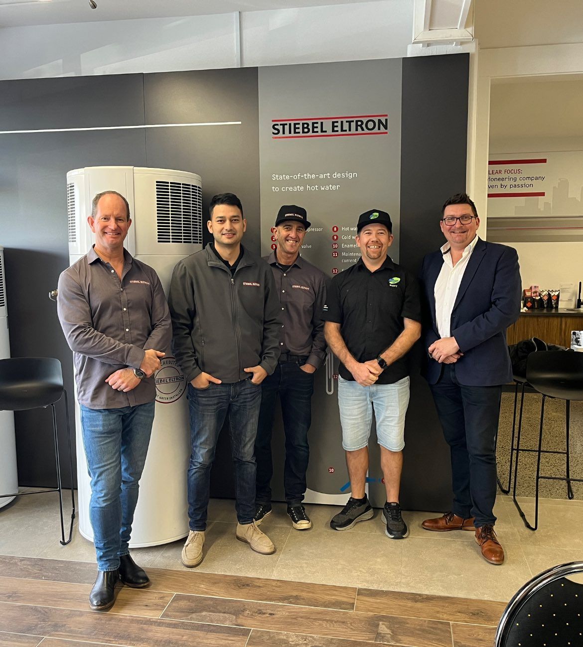 Five men standing near a Stiebel Eltron sign and water heater in an office.