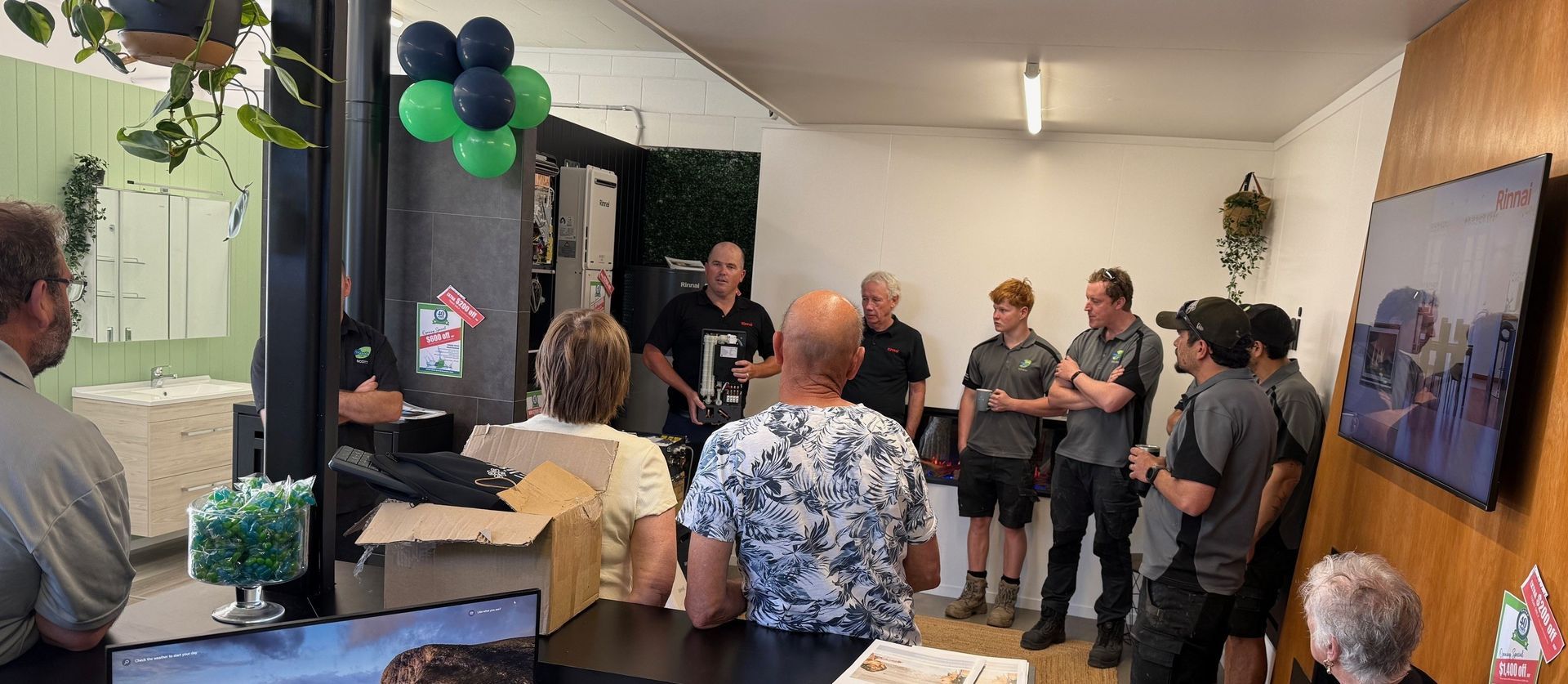 Group of people in a room, some listening to a speaker, others standing nearby. Balloons and plants.