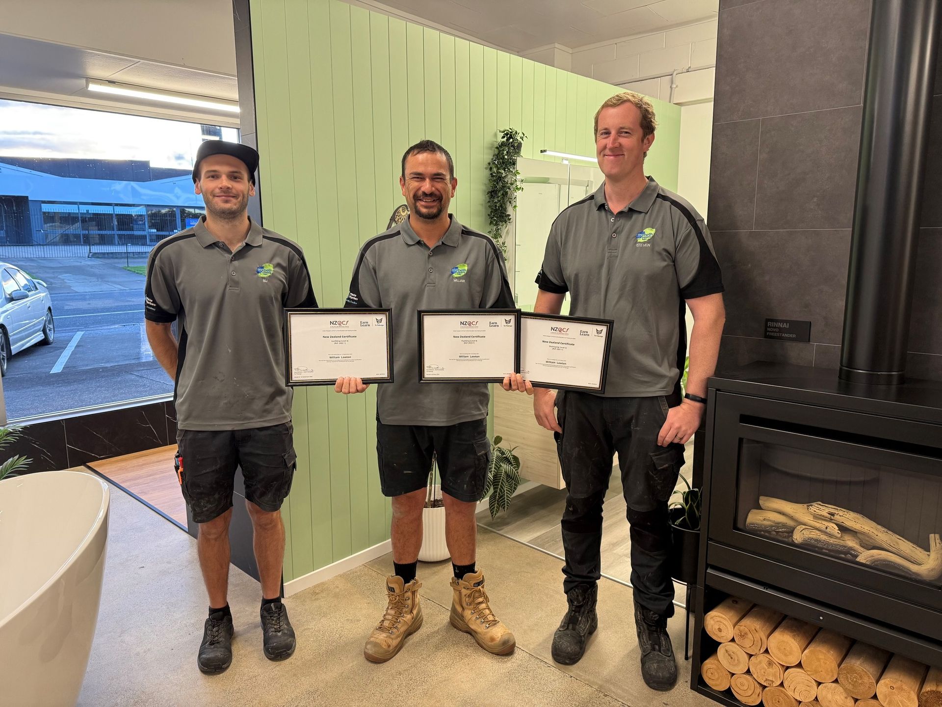 Three men holding certificates, smiling in a showroom with a fireplace and a bathtub.