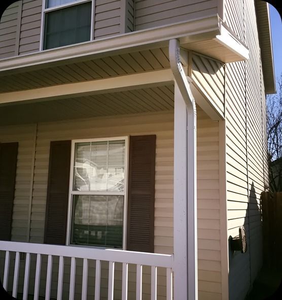 Tan house with white porch railing, brown shutters, and white gutters.
