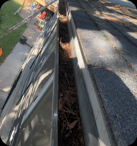 Gutter filled with debris, next to a dark roof, viewed from above.