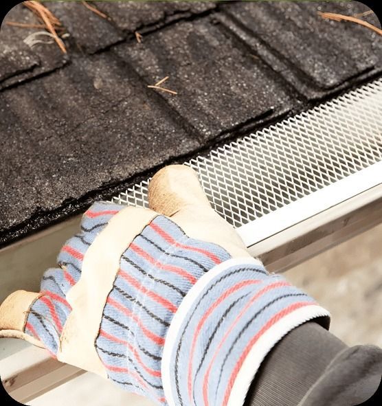 Gloved hand inspecting a gutter with mesh guard, next to a shingled roof.