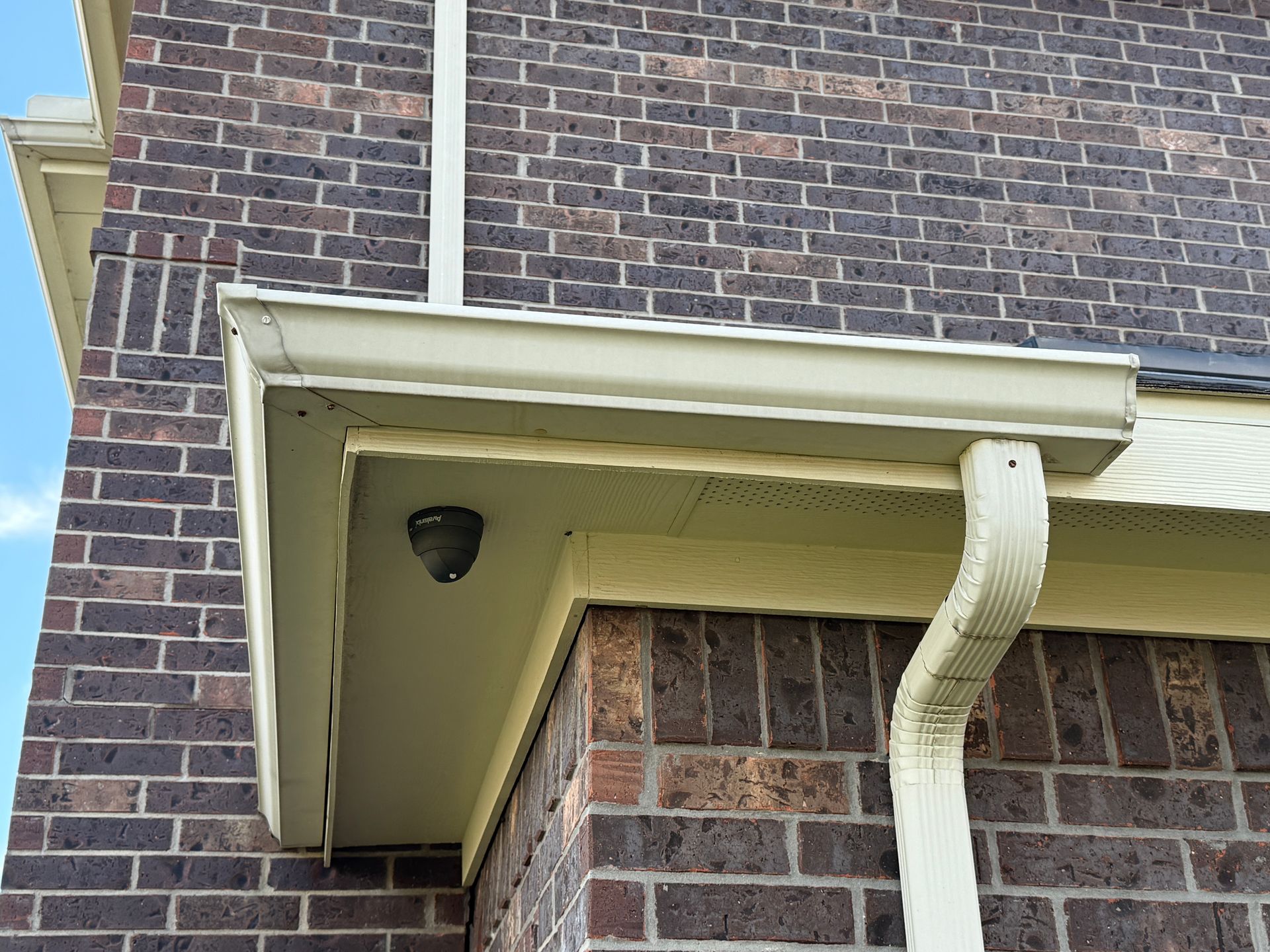 Exterior of a brick building with beige trim, a downspout, and a security camera under the eaves.