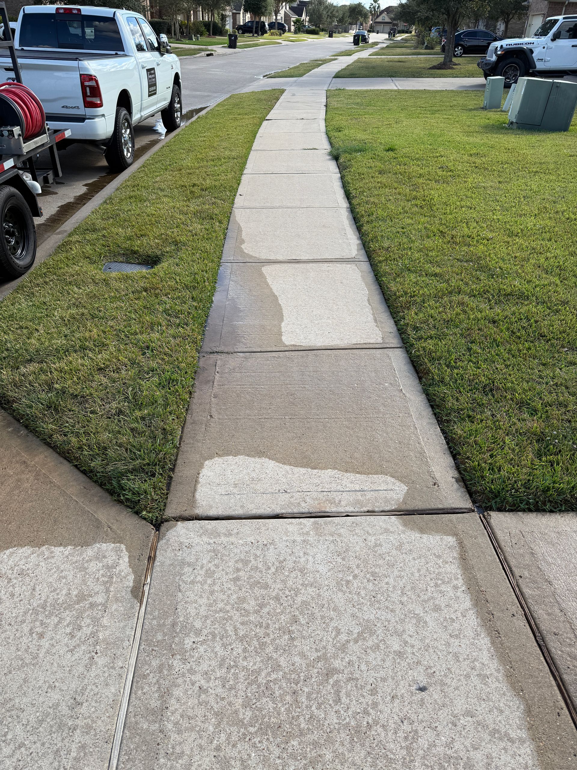 Sidewalk with wet spots, flanked by green grass and a street with a white pickup truck parked nearby.
