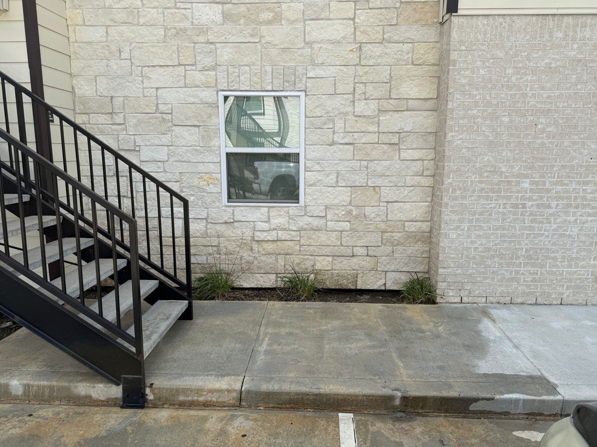 Exterior view of a building with stone facade, stairs, and a window. Small plants at the base.