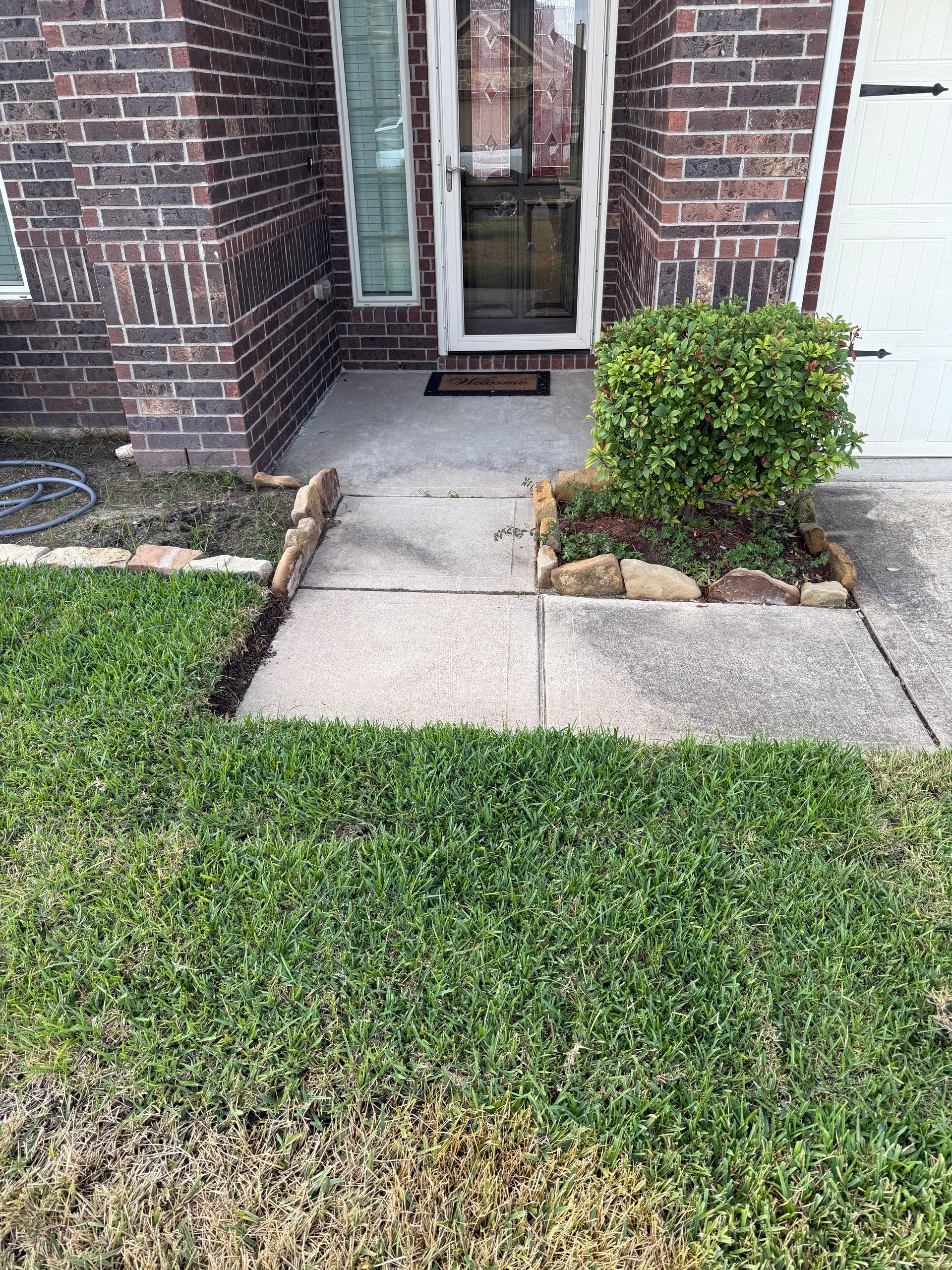 Concrete walkway to a front door with a small bush and grass. Brick building.