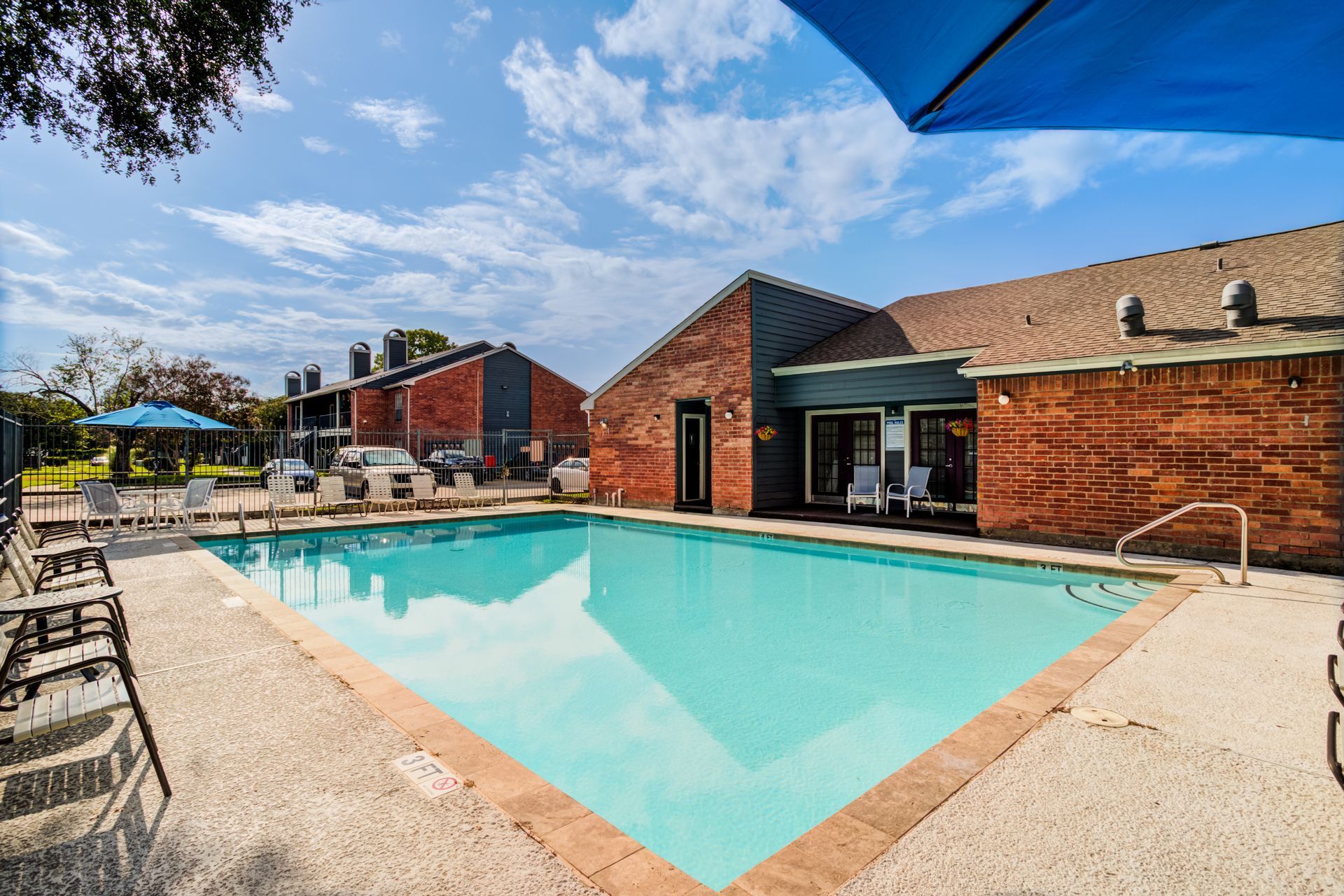 Pool area at apartments with brick building and blue umbrellas on a sunny day.