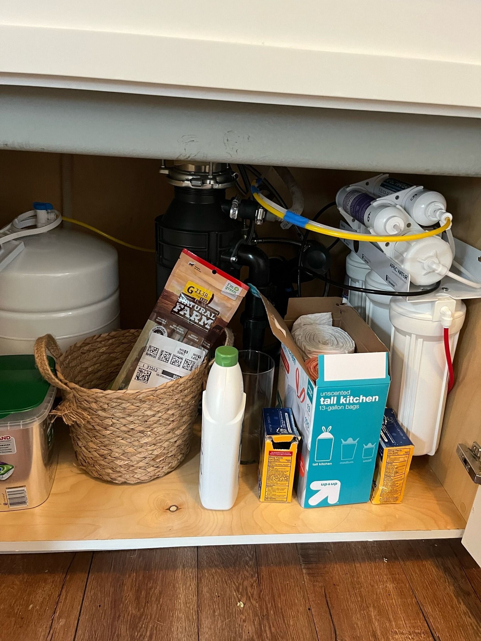 Under-sink area containing a white water tank, a filtration system, a woven basket, and various household cleaning supplies.