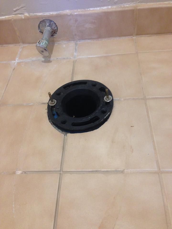 A black plastic toilet flange with two metal bolts installed on a beige tiled bathroom floor near a water supply line.