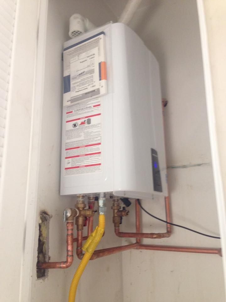 A white tankless water heater mounted in a small closet, connected to copper pipes and a yellow gas line.
