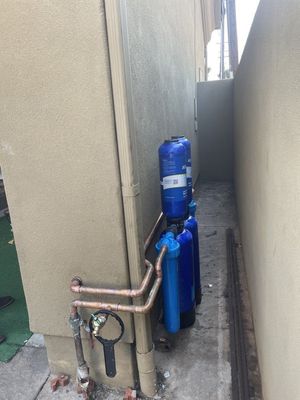 A side-yard alley containing a multi-stage blue water filtration system connected to copper pipes on a stucco wall.