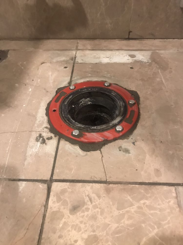 A toilet floor flange with a red metal ring secured to a tiled floor over an open drain pipe.