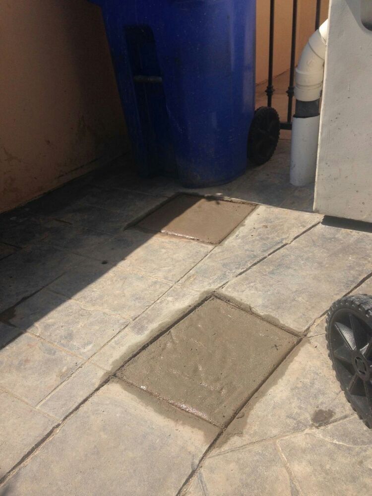 Two square patches of fresh concrete fill cutouts in a patio floor near a blue recycling bin and a white downspout.