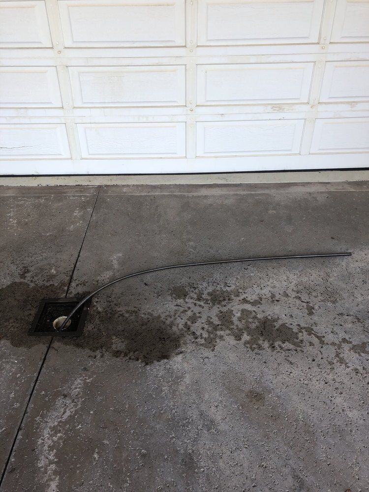 A coiled plumbing snake extending from a floor drain on a concrete driveway in front of a closed garage door.