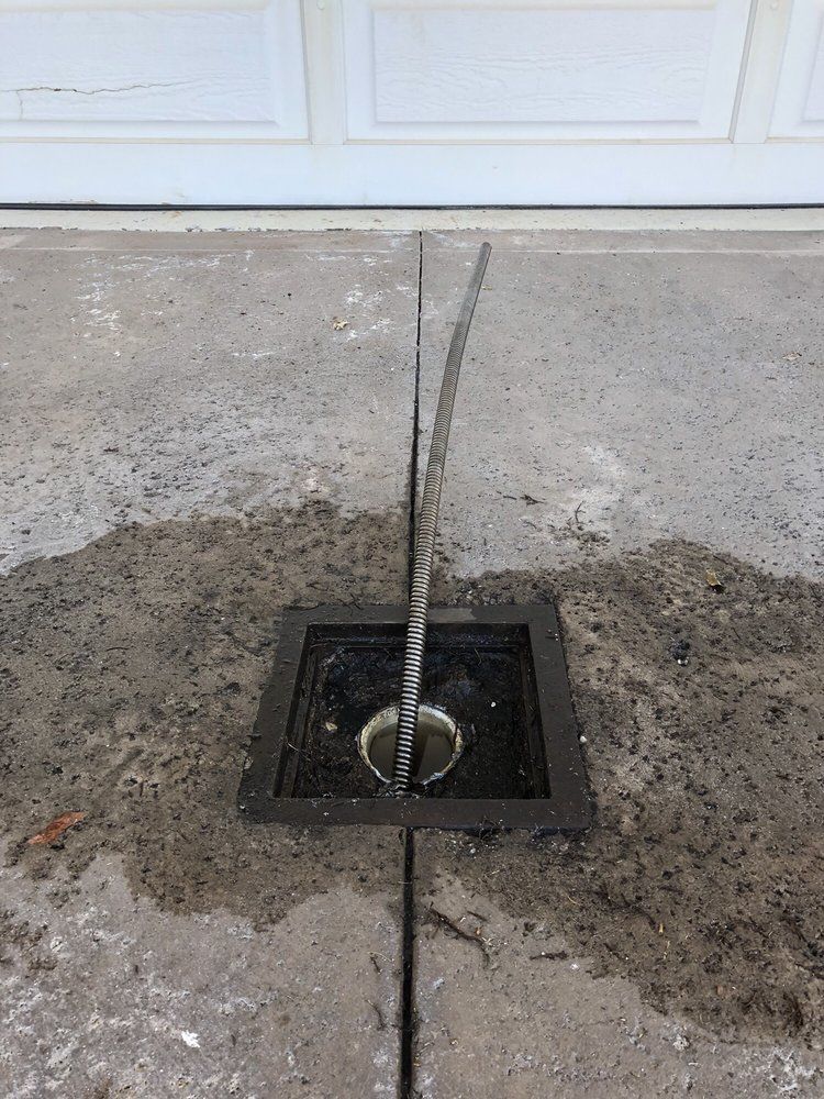A metal rod protrudes from a square concrete drain set in a driveway, bisected by a expansion joint leading to a garage.