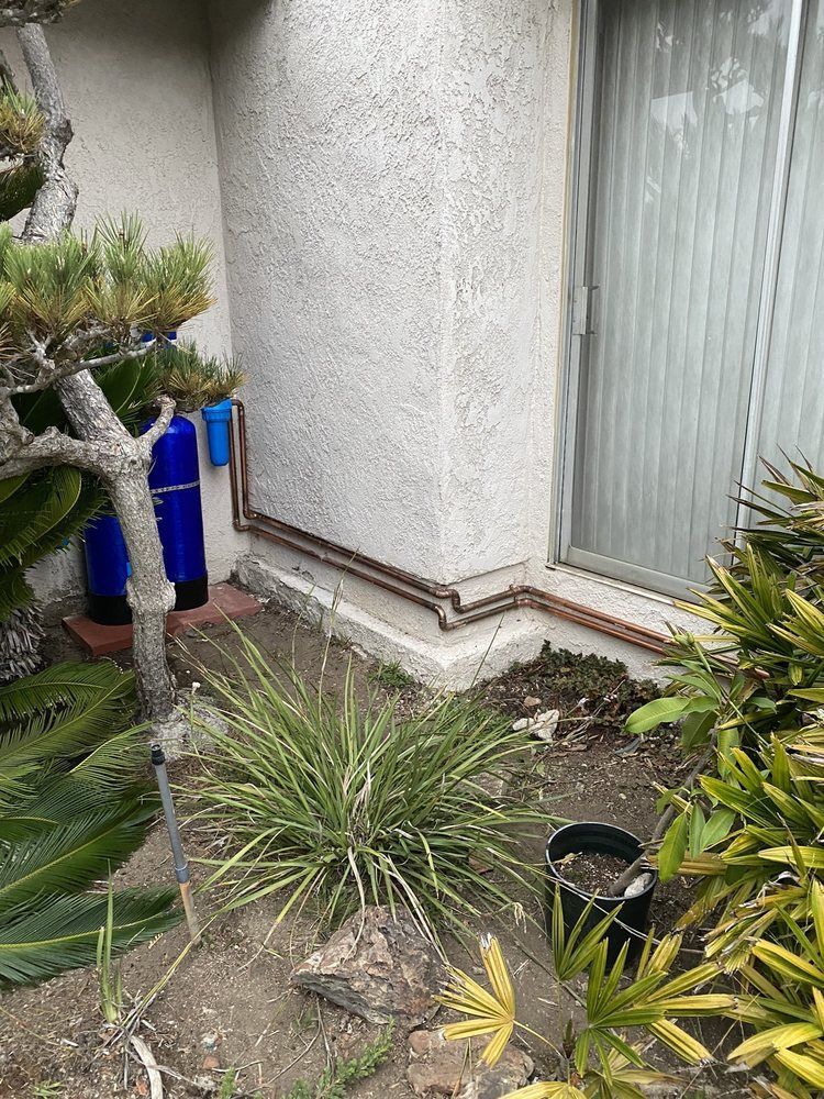 Blue water treatment tanks stand against the side of a house next to pipes and various plants in a dirt garden area.