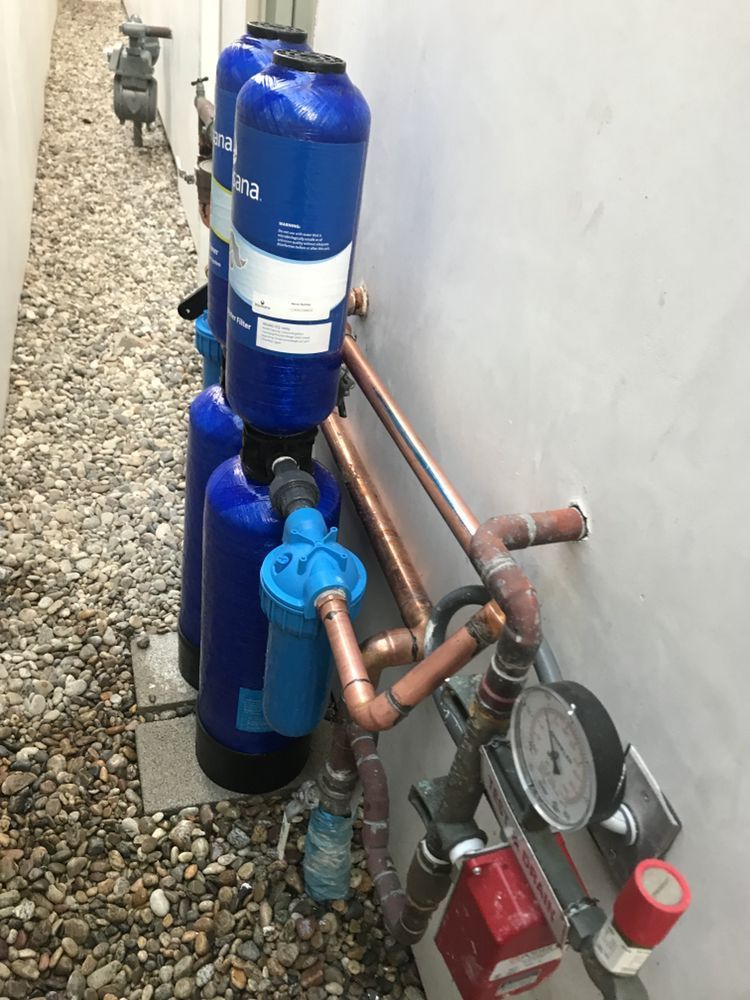 Two blue water filtration tanks installed on a gravel exterior wall next to copper plumbing pipes and a pressure gauge.