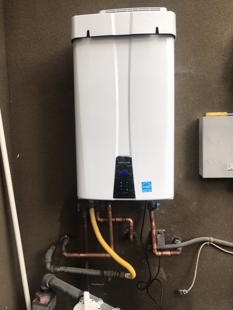 A white wall-mounted tankless water heater with copper plumbing connections, installed against a textured gray wall.